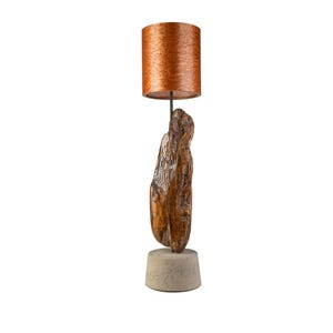 Op de afbeelding: Tafellamp met een koperkleurige, cilindrische lampenkap. De lamp heeft een betonnen voet en een stuk getextureerd hout. De lamp is ontworpen voor gebruik binnenshuis en heeft een moderne, rustieke uitstraling. De lamp is ongeveer 40 cm hoog.