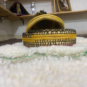 May include: A woven basket with a lid, featuring a yellow and black design. The basket is open, revealing its interior. It sits on a white, textured rug with green and brown accents. The lid has a black tassel.