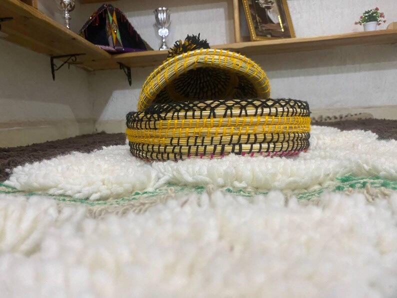 May include: A woven basket with a lid, featuring a yellow and black design. The basket is open, revealing its interior. It sits on a white, textured rug with green and brown accents. The lid has a black tassel.