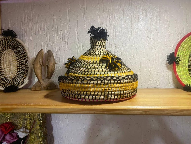 May include: A woven basket with a conical lid, featuring black and yellow patterns and black tassel accents. The basket sits on a wooden shelf, alongside other decorative items.