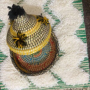 May include: A woven basket with a lid, featuring a black and yellow checkered pattern. The basket is adorned with black and yellow tassels. The interior of the basket is a colorful array of blue, red, and yellow. The basket is placed on a white rug with green stripes.