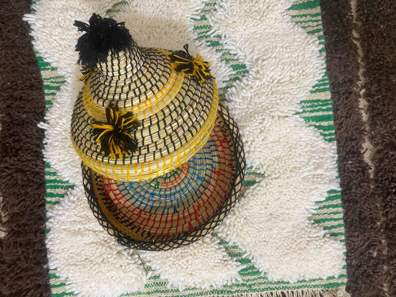 May include: A woven basket with a lid, featuring a black and yellow checkered pattern. The basket is adorned with black and yellow tassels. The interior of the basket is a colorful array of blue, red, and yellow. The basket is placed on a white rug with green stripes.