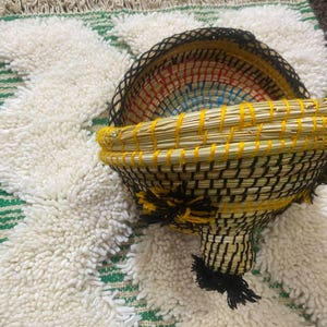 May include: A handcrafted, woven basket with a unique shape, featuring a yellow rim and black and yellow woven body. The interior displays a colorful pattern. The basket rests on a textured, white and green rug.