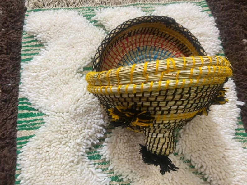May include: A handcrafted, woven basket with a unique shape, featuring a yellow rim and black and yellow woven body. The interior displays a colorful pattern. The basket rests on a textured, white and green rug.