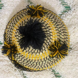 May include: A round, woven basket with a black and yellow pattern. The basket has a central black pom-pom and several smaller yellow and black pom-poms around the edge. The basket is sitting on a white and green patterned rug.