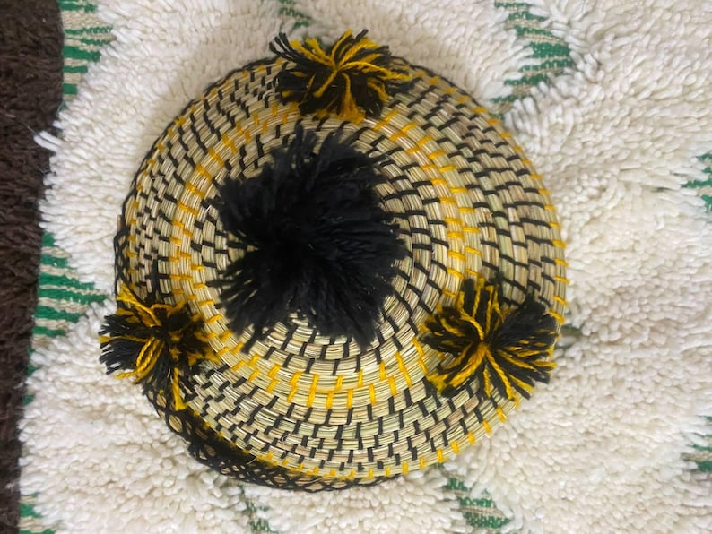 May include: A round, woven basket with a black and yellow pattern. The basket has a central black pom-pom and several smaller yellow and black pom-poms around the edge. The basket is sitting on a white and green patterned rug.