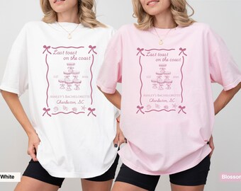 May include: White and pink t-shirts with a graphic design. The design includes the text "Last toast on the coast", "Ashley's Bachelorette Charleston, SC", and the year "2025". The design also features bows, seashells, and a tiered dessert.