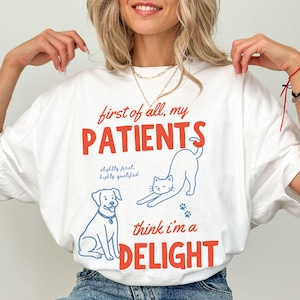 May include: White t-shirt with the text "first of all, my PATIENTS think I'm a DELIGHT" in red and blue. The shirt features illustrations of a dog, a cat, and paw prints. The text "slightly feral, highly qualified" is also included.