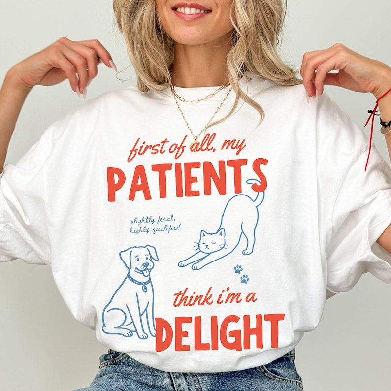 May include: White t-shirt with the text "first of all, my PATIENTS think I'm a DELIGHT" in red and blue. The shirt features illustrations of a dog, a cat, and paw prints. The text "slightly feral, highly qualified" is also included.
