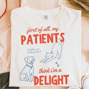 May include: Cream-colored sweatshirt with the text "first of all, my PATIENTS" in red, and "think i'm a DELIGHT" in red. The design includes a blue dog and cat illustration, with the words "slightly feral, highly qualified" in blue.