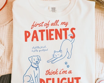 May include: Cream-colored sweatshirt with the text "first of all, my PATIENTS" in red, and "think i'm a DELIGHT" in red. The design includes a blue dog and cat illustration, with the words "slightly feral, highly qualified" in blue.