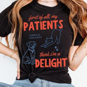May include: Black t-shirt with red and blue graphic text and illustrations. The text reads "first of all, my PATIENTS" and "think i'm a DELIGHT". Illustrations include a dog, a cat, and paw prints.