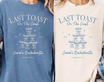 May include: Two t-shirts, one blue and one cream, with the text "LAST TOAST On The Coast" and "Sarah's Bachelorette" in blue script. The design includes a champagne tower illustration and the year 2025.