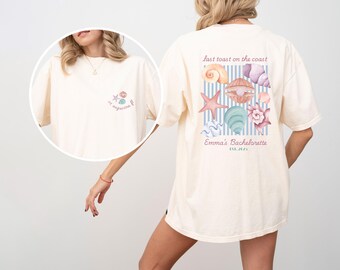 May include: Cream-colored t-shirt with a beach-themed design. The front features a small graphic with a starfish and seashell. The back displays various seashells, a starfish, and the text "last toast on the coast" and "Emma's Bachelorette".
