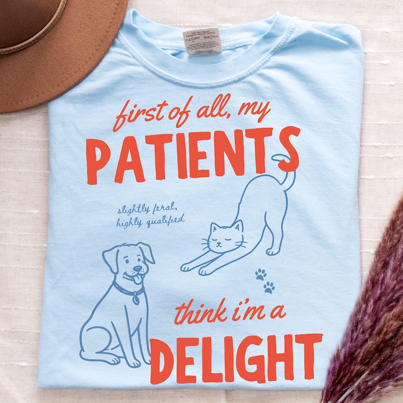 May include: Light blue t-shirt with the text "first of all, my PATIENTS think I'm a DELIGHT." The shirt features illustrations of a dog and a stretching cat. The text "slightly feral, highly qualified" is also printed on the shirt.