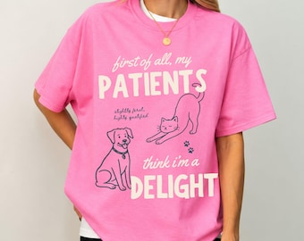 May include: A bright pink short-sleeved t-shirt featuring a dark blue graphic of a dog and a stretching cat. The text reads: first of all, my PATIENTS, slightly feral, highly qualified, think i'm a DELIGHT. A gold necklace is worn around the neck.