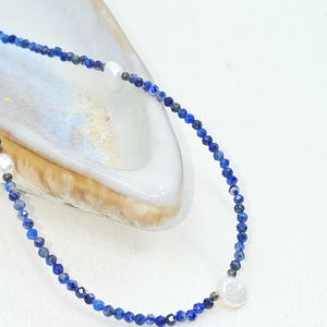 May include: A delicate necklace featuring small, faceted blue beads and three white, round pearls. The necklace is displayed on a white surface, showcasing its elegant design and color contrast. The necklace is a piece of jewelry.
