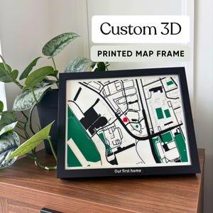 Può includere: Una mappa stampata in 3D con cornice nera, caratterizzata da sezioni bianche, nere e verdi e un piccolo segnaposto rosso. La cornice include una targhetta con il testo "Our first home" in basso. La mappa mostra un layout stradale stilizzato.