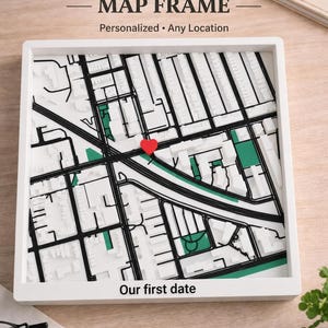 May include: A white framed map with a red heart marking a location. The map is a detailed street view with green and black accents. Text at the top reads "Custom Location MAP FRAME" and "Personalized • Any Location." The bottom text says "Our first date."