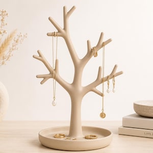 May include: A beige, tree-shaped jewellery stand with a circular base. The stand features multiple branches for hanging necklaces and earrings. Several gold-toned necklaces with pendants, a ring, and hoop earrings are displayed on the branches and base.