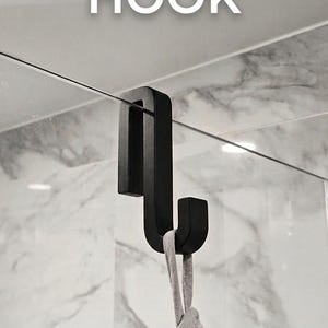 Peut inclure: Un crochet de douche en métal noir mat conçu pour être suspendu sur une paroi en verre. Le crochet présente une section supérieure rectangulaire qui s'adapte au bord du verre et un crochet inférieur courbé retenant une serviette grise.