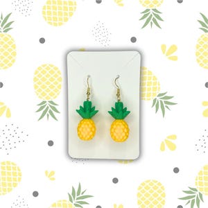 Peut inclure: Une paire de boucles d'oreilles ananas jaunes avec des dessus verts et des crochets dorés. Les boucles d'oreilles sont présentées sur une carte blanche sur un fond blanc avec un motif d'ananas.