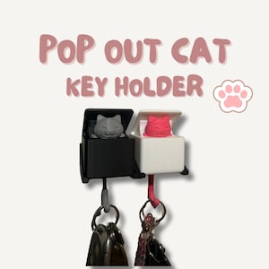 Puede incluir: Dos llaveros con temática de gato, uno negro y otro blanco, cada uno con un diseño de gato desplegable. Los llaveros tienen un llavero adjunto. El texto "POP OUT CAT KEY HOLDER" se muestra encima de los llaveros.