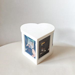May include: A white, heart-shaped photo cube with printed photos visible on three sides. The cube is made of wood and has a clean, modern design. The photos appear to be personal snapshots.