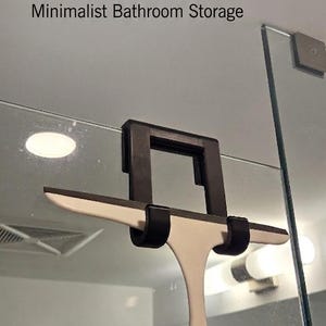 May include: A shower squeegee holder with the text "SHOWER SQUEEGEE HOLDER Minimalist Bathroom Storage". The holder is black and white, designed to hold a squeegee. The squeegee is white with a black handle.