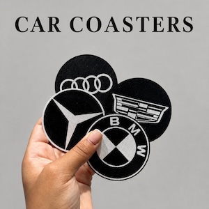 May include: A set of four circular black car coasters featuring white logos for Audi, Mercedes-Benz, Cadillac, and BMW. The text CAR COASTERS is printed in black above the coasters. Each coaster has a distinct automotive brand emblem design.