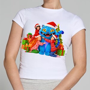 K&ouml;nnte beinhalten: Wei&szlig;es T-Shirt mit einem weihnachtlichen Motiv. Das Design zeigt eine blaue Katze mit Weihnachtsm&uuml;tze, umgeben von Kindern und Geschenken. Ein Weihnachtsbaum ist ebenfalls im Hintergrund zu sehen.