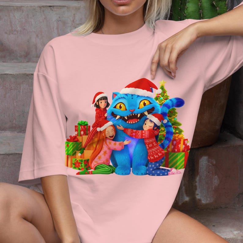 K&ouml;nnte beinhalten: Hellrosa &uuml;bergro&szlig;es T-Shirt mit einem weihnachtlichen Motiv. Die Grafik zeigt eine blaue Katze mit Weihnachtsm&uuml;tze, die von drei Kindern umarmt wird. Weihnachtsgeschenke und ein Weihnachtsbaum sind ebenfalls im Design zu sehen.