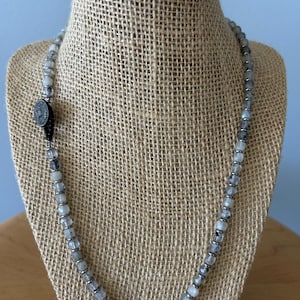 May include: A beaded necklace featuring small, faceted, translucent gray and white stones interspersed with tiny metallic silver spacers. A dark, circular, textured charm is attached to the left side of the strand, secured by a black cord.