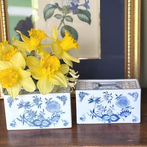 May include: Two white and blue floral ceramic vases with yellow daffodils. The vases have a rectangular shape and a blue floral pattern. The background includes a framed botanical print.