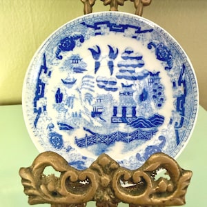 May include: A round ceramic saucer featuring a blue and white willow pattern. The design depicts a landscape with pagodas, trees, a bridge, and two birds in flight. The saucer is displayed on an ornate, metallic, bronze-colored stand.