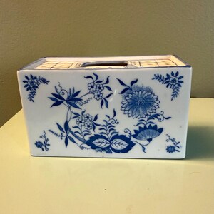 May include: A rectangular ceramic tissue box cover featuring a white base with a blue floral pattern. The design includes intricate botanical illustrations of flowers, leaves, and vines across the front and top surfaces, with a central opening.