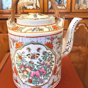 Puede incluir: Tetera decorativa de porcelana con asa trenzada. La tetera presenta un diseño floral y de mariposas en tonos rosa, naranja y verde. El pico es blanco con un patrón decorativo. La tetera mide aproximadamente 20 cm de alto.