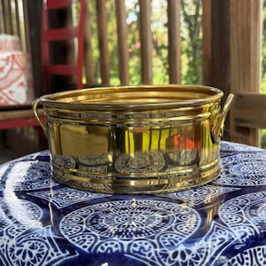 Puede incluir: Un pequeño macetero redondo de latón con dos asas. El macetero está sobre una superficie con un patrón azul y blanco.