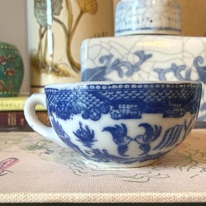 May include: A white ceramic teacup featuring a classic blue willow pattern. The design displays intricate illustrations of two birds facing each other, architectural structures, and geometric borders encircling the exterior of the cup.