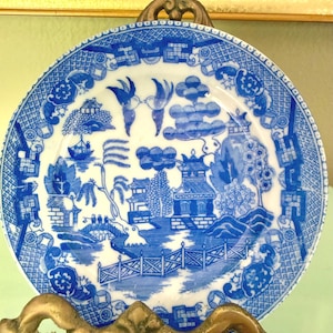 May include: A round ceramic plate featuring a blue and white willow pattern. The design depicts a landscape with a bridge, pagodas, trees, and two birds flying at the top. The plate is displayed on a decorative metal stand with ornate details.