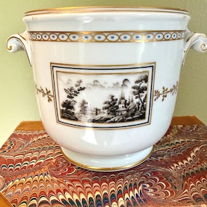 May include: A white porcelain cachepot featuring two side handles, a gold rim, and a decorative band with blue dots. The center displays a rectangular black and white landscape illustration of trees and buildings, accented by gold leaf vine motifs.
