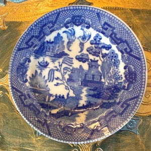 May include: A round, blue and white ceramic plate with a Willow pattern design. The plate features a detailed landscape scene with bridges, buildings, and trees. The rim has a decorative border.