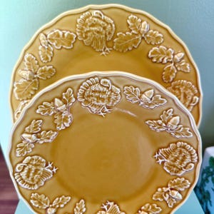 May include: Two round, mustard-yellow ceramic plates featuring a scalloped edge and a raised, embossed border design. The border pattern consists of alternating turkey figures and oak leaves with acorns, creating a textured, relief-style surface.