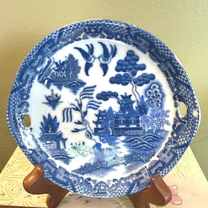 May include: A blue and white ceramic plate with a Willow pattern design. The plate features a detailed landscape scene with pagodas, trees, and a bridge. Two small handles are on either side of the plate. The plate is displayed on a wooden stand.