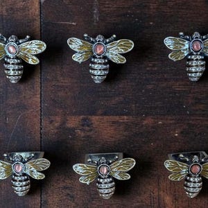 May include: Ten silver-toned bee-shaped rings arranged in two rows. Each bee features gold-colored wings, a small round pink stone at the center of the thorax, and a striped abdomen with clear crystal accents along the body and wings.
