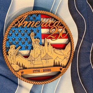 May include: A circular wooden plaque with a layered design. It features the word "America" and "250", alongside the American flag, the Statue of Liberty, a soldier, and an eagle. The years 1776-2026 are also displayed on the plaque.
