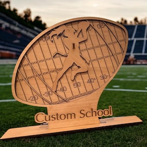 May include: A wooden football-shaped decorative piece with a football field design. A cutout of a football player is running with the ball. The text "Custom School" is at the base. The piece is made of light-colored wood.