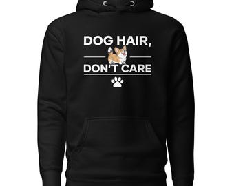 以下が含まれることがあります： DOG HAIR, DON'T CAREという白いグラフィックプリントが施された黒いフード付きスウェットシャツ。2行のテキストの間には、茶色と白のコーギー犬の小さなイラストが描かれています。テキストの下には白い肉球のアイコンがあります。