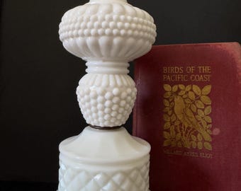Puede incluir: Una lámpara de mesa de vidrio blanco con un diseño decorativo. La lámpara tiene un cuerpo texturizado y bulboso y una base con patrón de diamantes. Un libro rojo titulado "Birds of the Pacific Coast" está al lado de la lámpara.