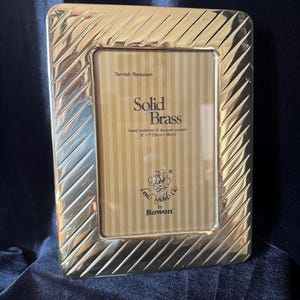 May include: A rectangular, gold-colored brass picture frame with a diagonal grooved pattern around the edges. The frame holds a striped, tan-colored backing. Text on the frame reads "Solid Brass" and "5" x 7" (13cm x 18cm)".